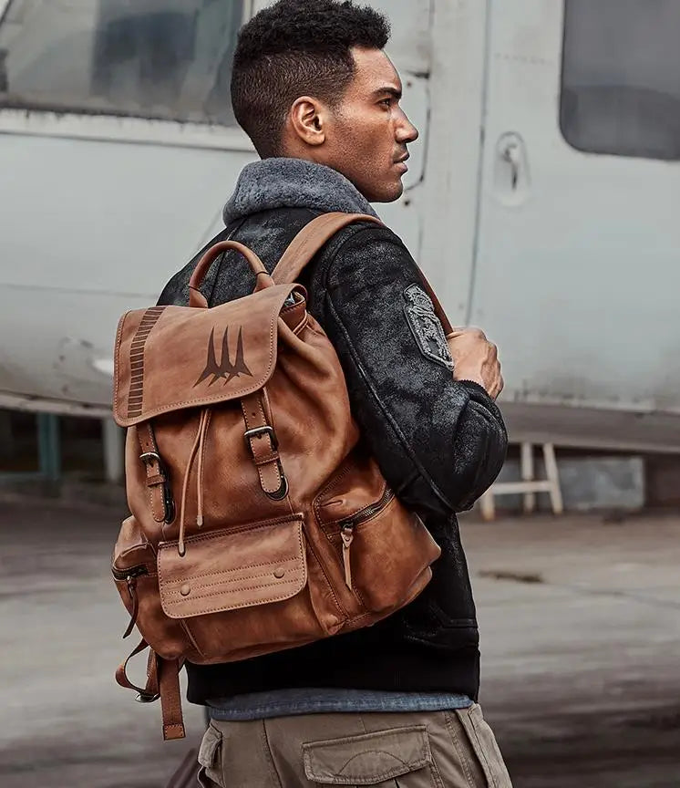 A man wearing a brown distressed leather backpack with a black triangular logo on the front flap, styled with a black jacket and khaki pants, standing in front of a gray aircraft.