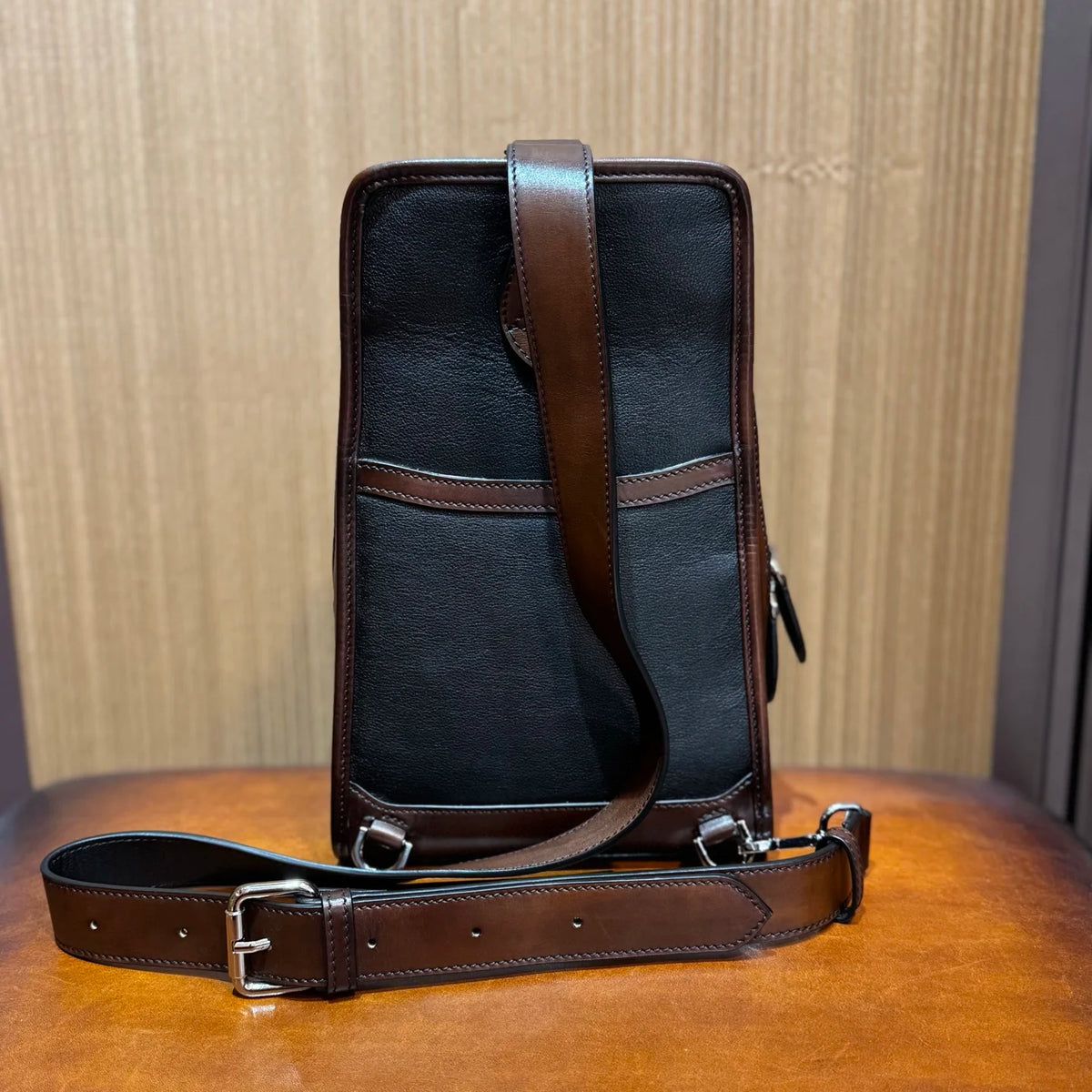 A black leather and canvas crossbody bag with dark brown leather trim and strap, displayed on a wooden surface against a wood-paneled background.
