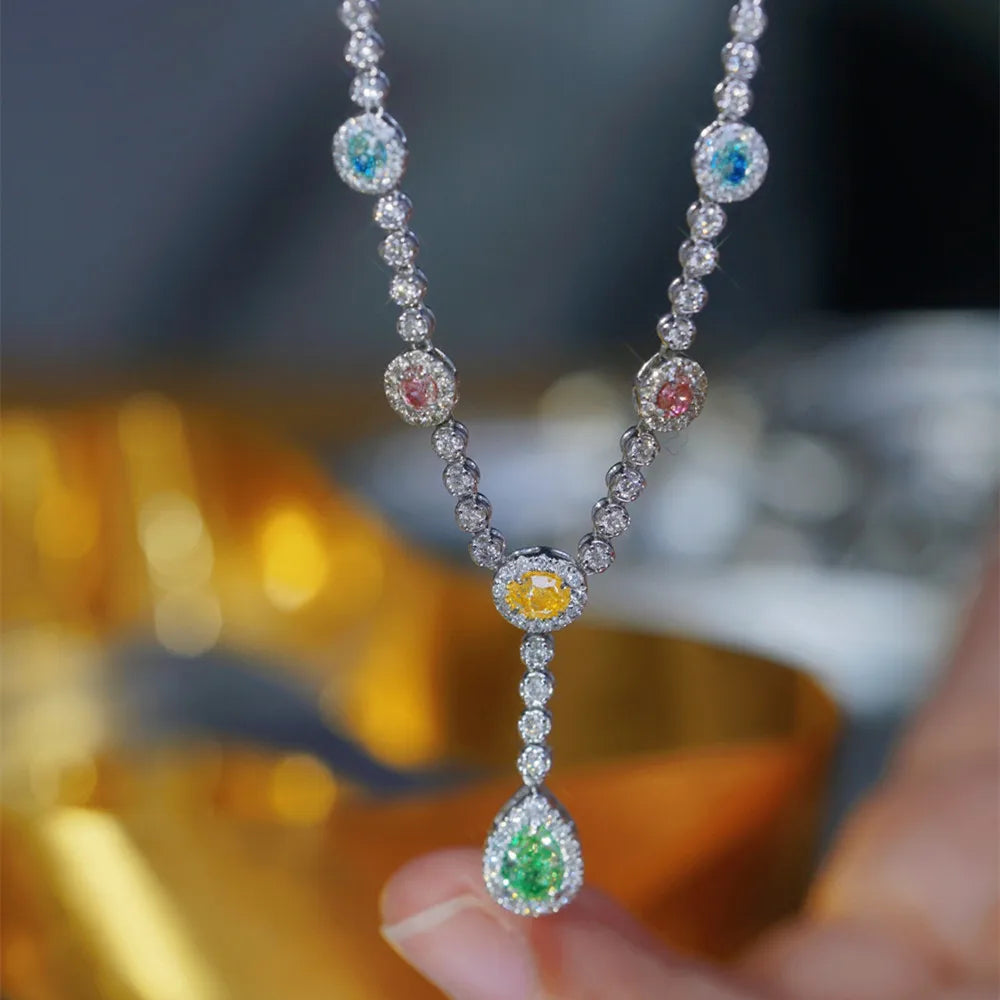 A close-up of a silver-toned Y-shaped necklace with round and teardrop-cut colored gemstones, held by fingers against a blurred golden and dark background.