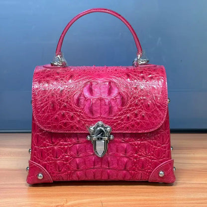 A bright pink crocodile-embossed leather handbag with a top handle, silver-tone hardware, and a decorative front clasp, photographed on a wooden surface against a blue wall.