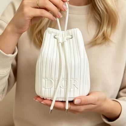 A person holding a small pleated white drawstring bucket bag with embossed Dior branding, wearing a beige knit sweater.