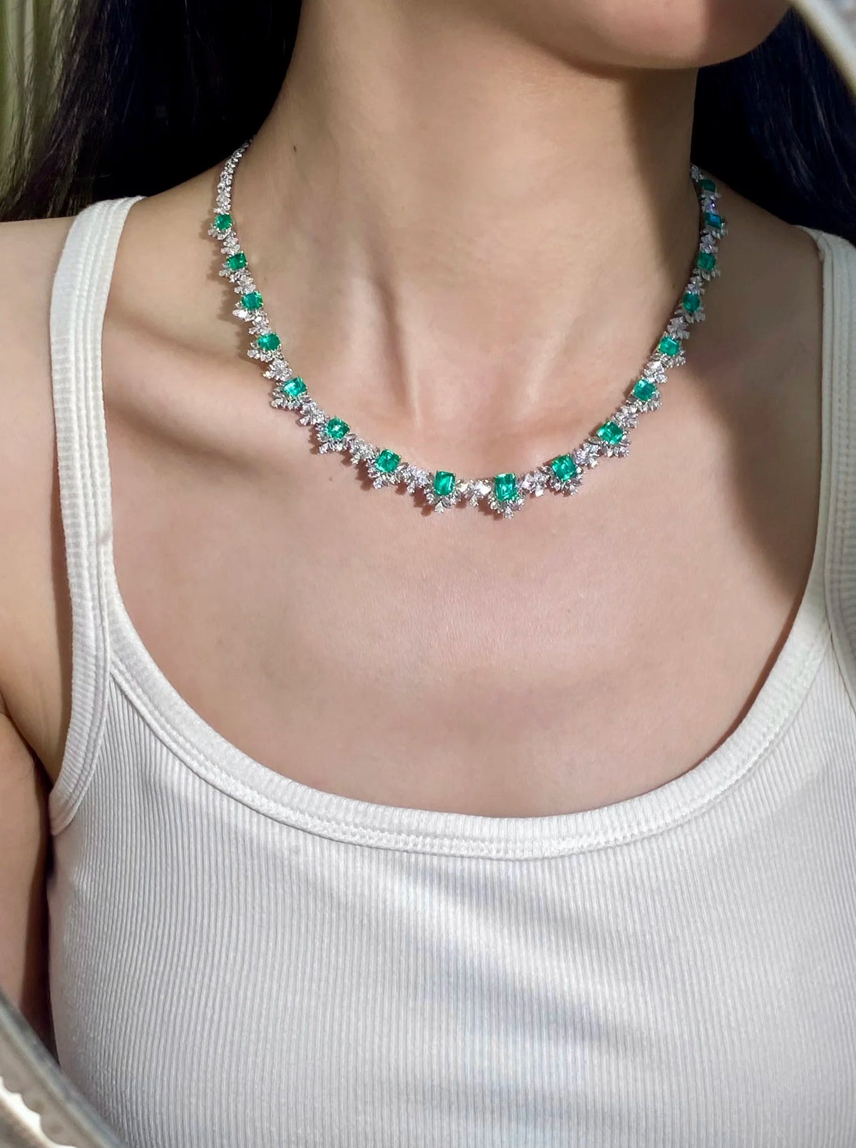 A close-up of a woman wearing a silver-toned necklace adorned with green emerald-cut gemstones and clear round stones, paired with a white ribbed tank top.