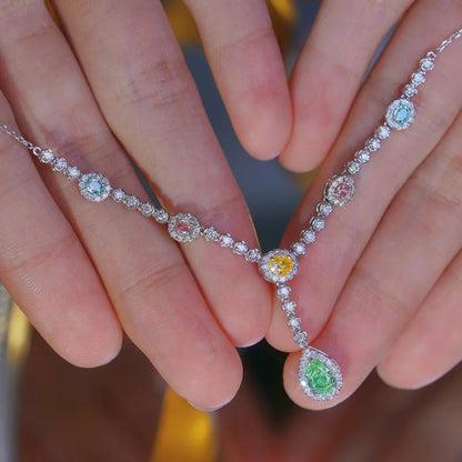 A close-up of a delicate silver Y-shaped necklace with colorful gemstones, held between two hands. The necklace features a central yellow gemstone and a larger green teardrop gemstone, surrounded by smaller pink, blue, and green stones on a silver chain.