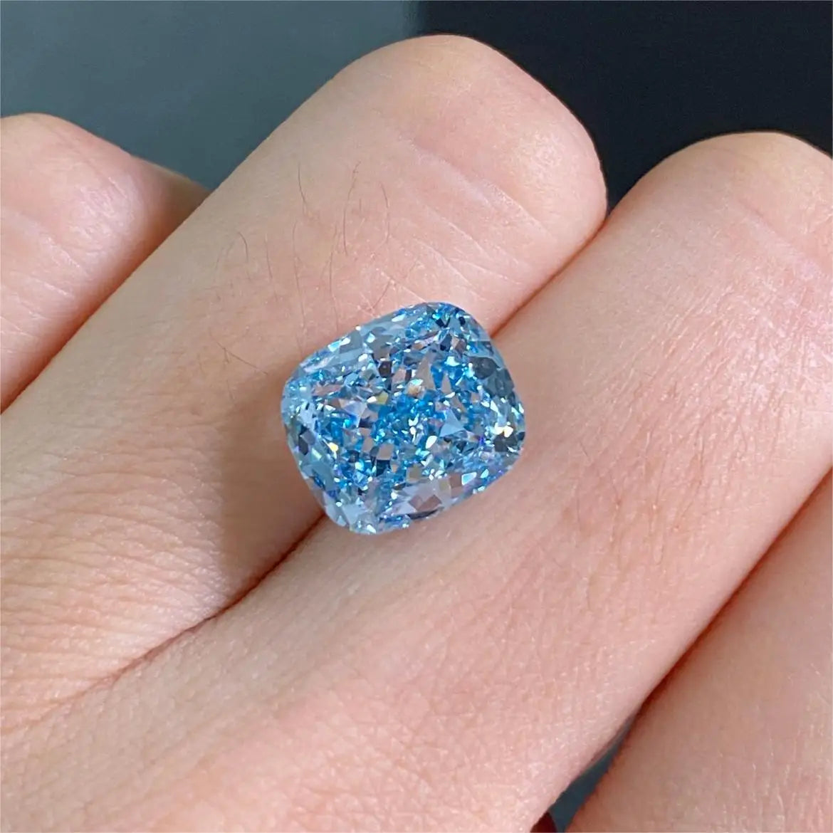 A loose cushion-cut light blue gemstone displayed on a person's finger against a dark gray background.