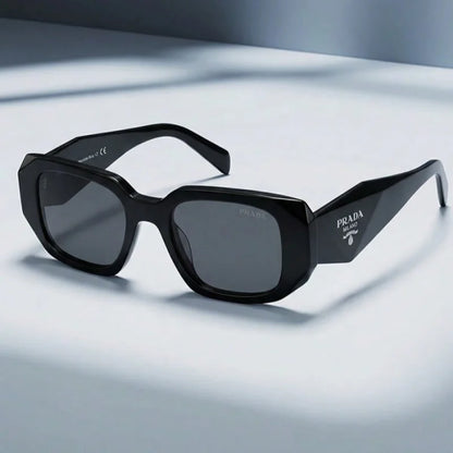 Prada sunglasses with a glossy black acetate frame, dark gray lenses, and the Prada logo printed on the temple arm, displayed on a clean light gray surface.
