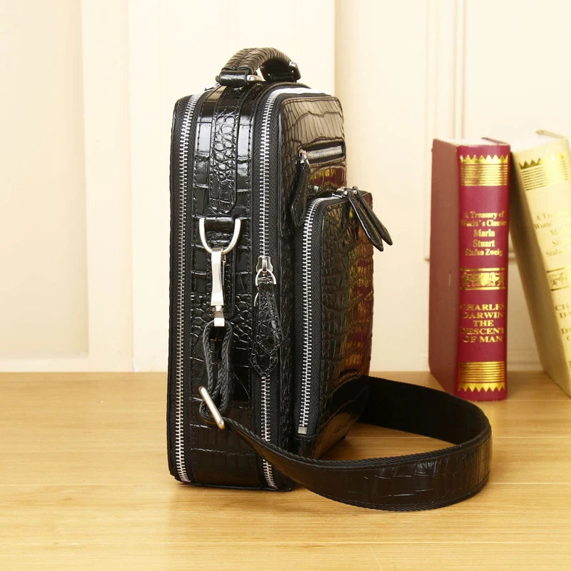 A black crocodile-embossed leather crossbody bag with silver zippers and a matching shoulder strap, standing on a wooden surface next to two books.