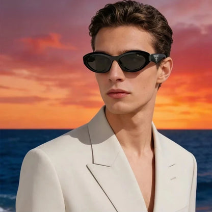 A man wearing black cat-eye sunglasses and a white blazer, set against a dramatic sunset over the ocean.
