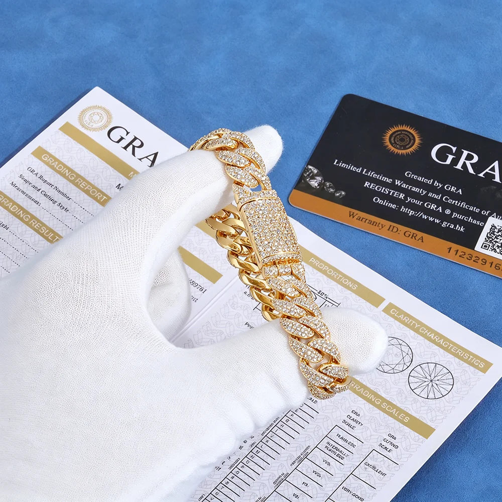 A gold-toned Cuban link chain bracelet covered in clear pavé stones is held by a white gloved hand over blue-gray papers and a black/grandfathered warranty card. The product is shown with documentation and a grandfathering card, indicating a luxury or pre-owned jewelry listing.