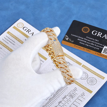 A gold-toned Cuban link chain bracelet covered in clear pavé stones is held by a white gloved hand over blue-gray papers and a black/grandfathered warranty card. The product is shown with documentation and a grandfathering card, indicating a luxury or pre-owned jewelry listing.