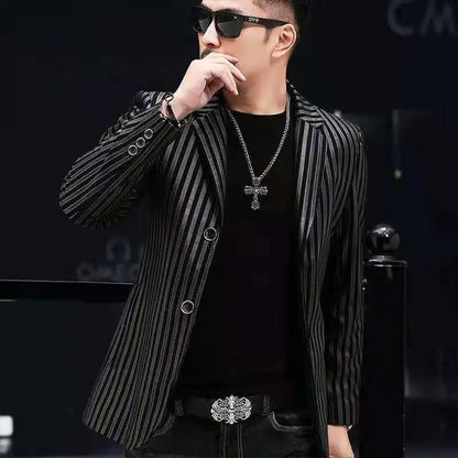 A man wearing a black and gray vertically striped blazer over a black t-shirt, styled with a silver cross necklace, sunglasses, and a patterned belt, posing in a dark setting.
