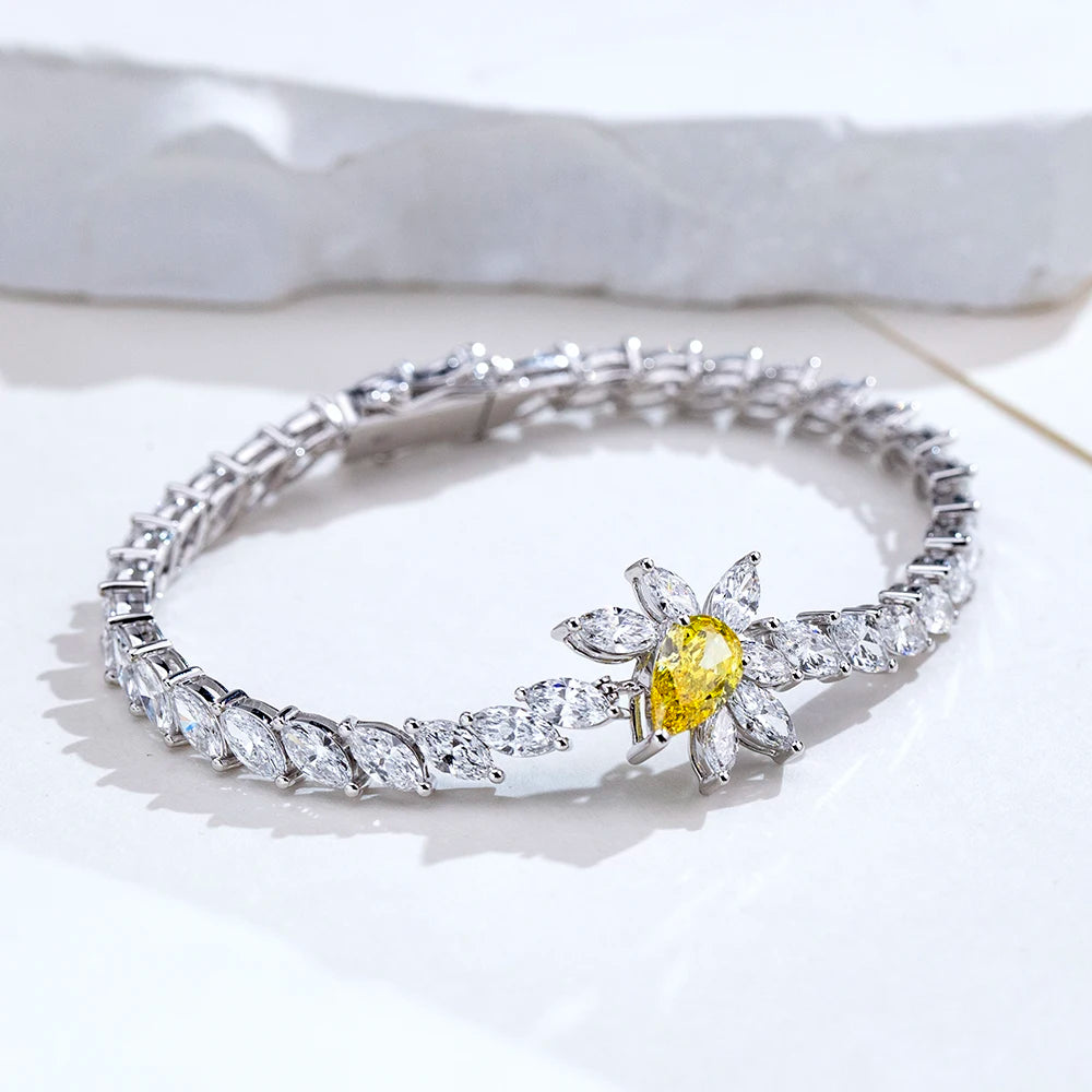 A silver-toned tennis bracelet featuring a central flower motif with a bright yellow oval center stone and surrounding clear marquise and round stones, displayed on a white surface.