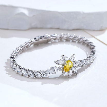 A silver-toned tennis bracelet featuring a central flower motif with a bright yellow oval center stone and surrounding clear marquise and round stones, displayed on a white surface.