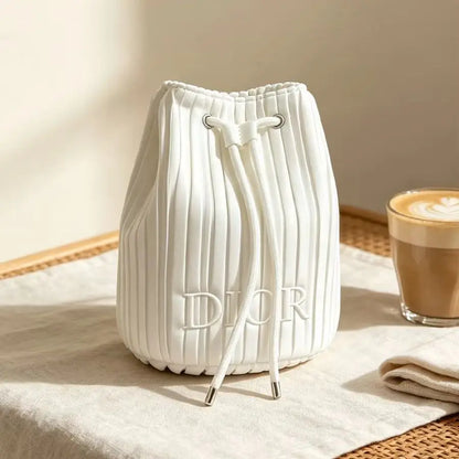 A white pleated drawstring bucket bag with embossed 'DIOR' lettering, styled on a beige fabric surface next to a cup of latte in a warm, sunlit setting.