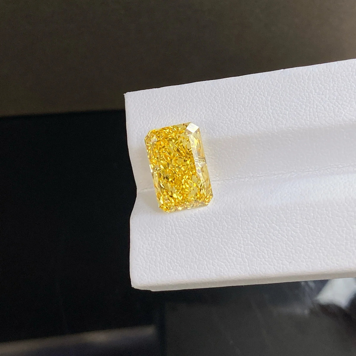 A rectangular yellow gemstone with a rough cushion cut, showing vibrant yellow color and sparkling facets, displayed on a white textured surface.