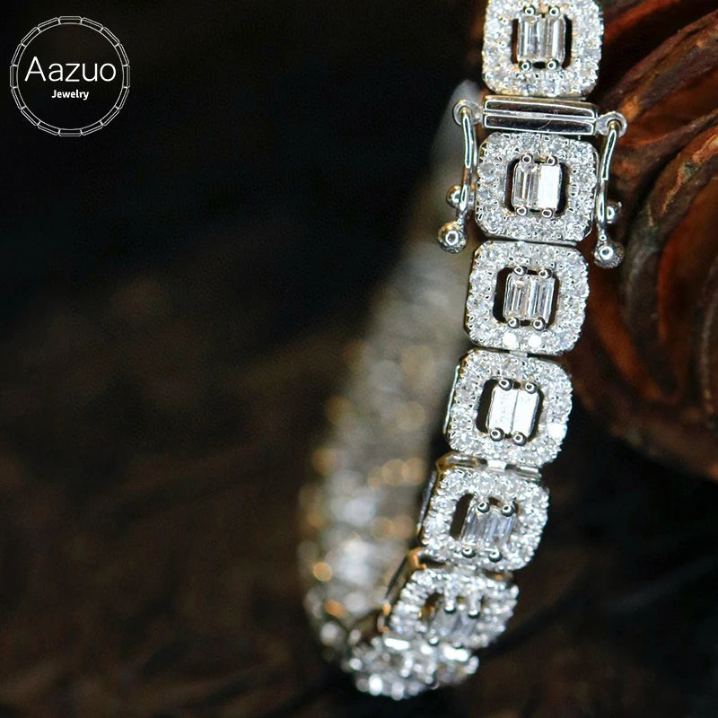 A close-up of a silver-toned tennis bracelet with baguette-cut clear stones, resting on a dark textured surface with a wooden prop. The Aazuo Jewelry logo is visible in the top-left corner.