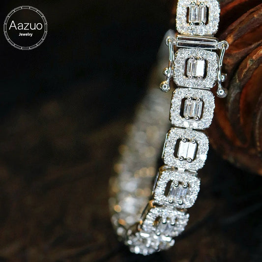 A close-up of a silver-toned tennis bracelet with baguette-cut clear stones, resting on a dark textured surface with a wooden prop. The Aazuo Jewelry logo is visible in the top-left corner.