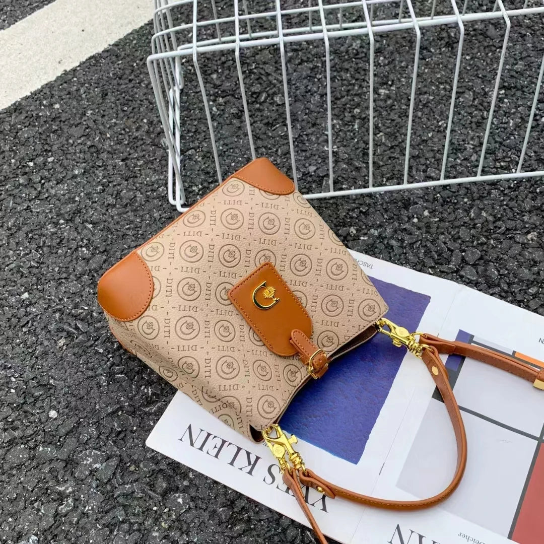 A beige monogram shoulder bag with tan leather trim and gold hardware, styled on top of a magazine over dark asphalt with a metal wire basket in the background.