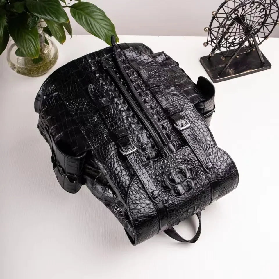 A black crocodile-embossed leather backpack styled on a white tabletop with a green plant in a glass vase on the left and a decorative metal wheel sculpture on the right.