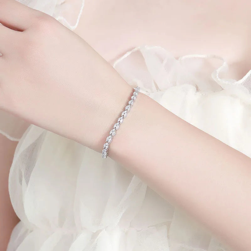 A delicate silver-toned tennis bracelet with clear stones, worn on a woman's wrist against a soft white tulle dress background.