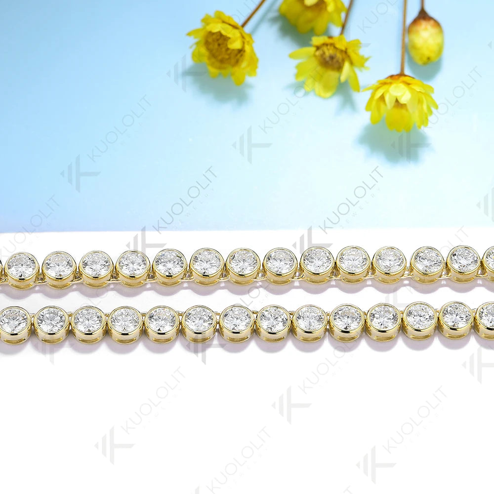 Two strands of round-cut clear gemstones in gold bezel settings, laid horizontally on a white surface with a light blue background and yellow flowers in the upper corner.