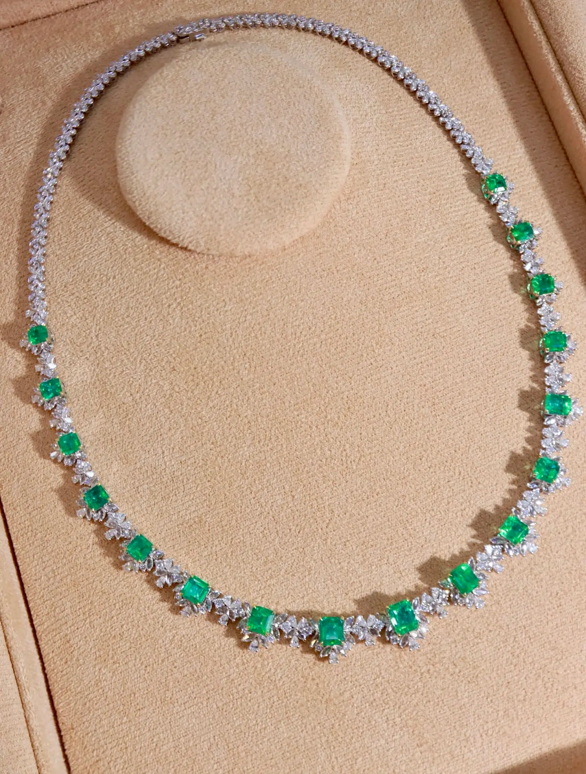 A diamond and emerald necklace displayed on a beige velvet jewelry stand. The necklace features a silver-toned chain with clusters of round-cut green emeralds and round brilliant-cut diamonds, arranged in a semi-circular shape on the display.