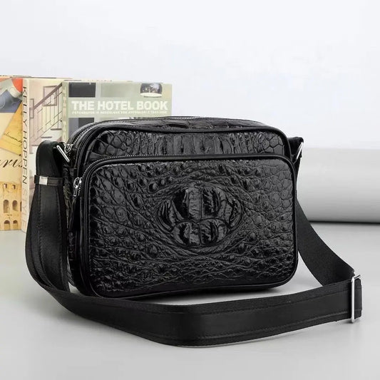 Black crocodile-embossed leather crossbody bag with a front zip pocket and adjustable shoulder strap, displayed on a light gray surface with books in the background.