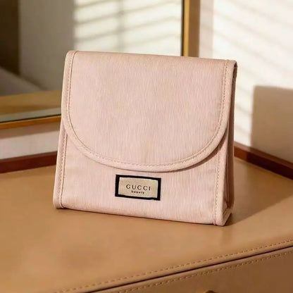 A light pink Gucci wallet made of textured canvas, featuring a black and beige logo patch on the front, resting on a tan leather surface with warm sunlight and window blinds in the background.