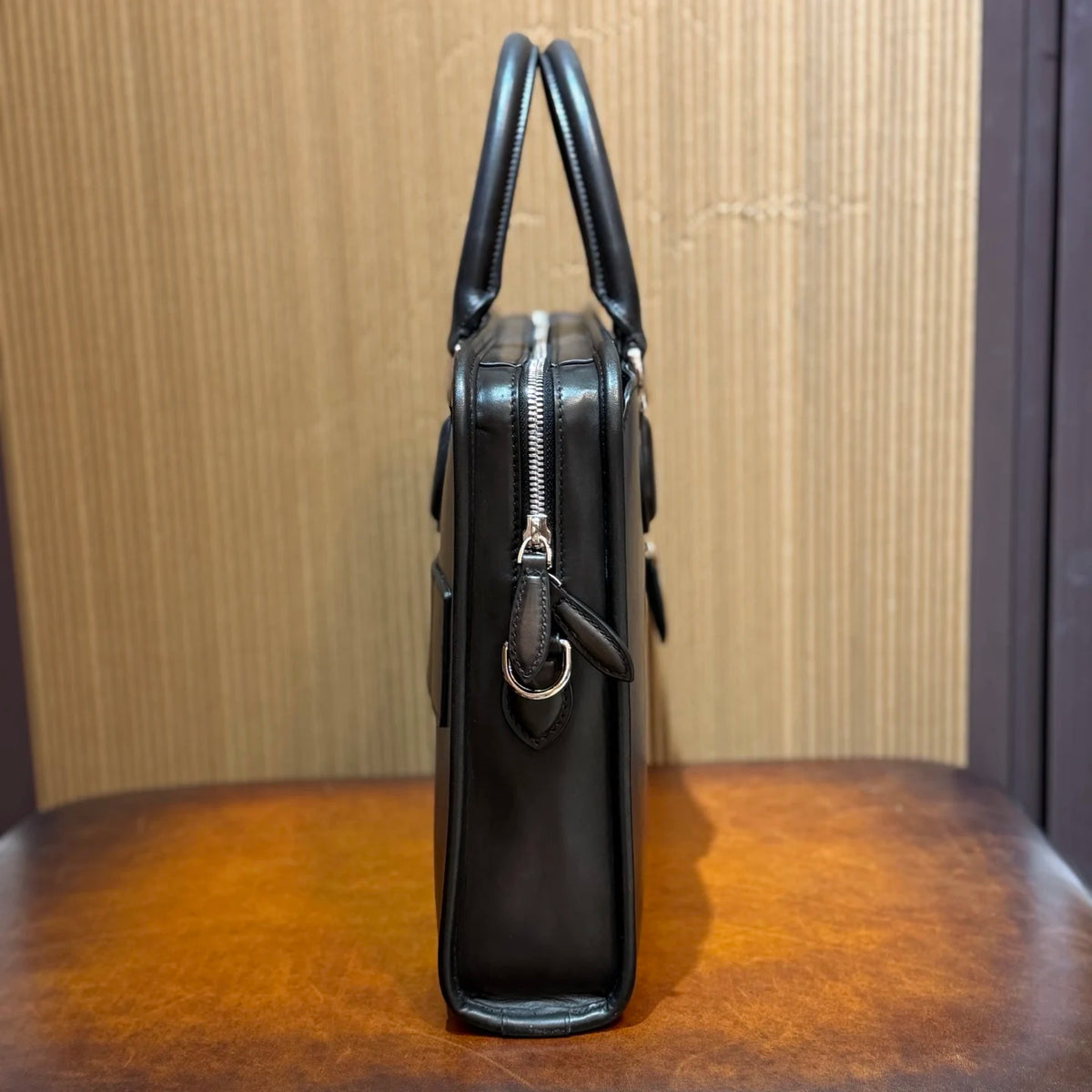 A black leather briefcase or satchel shown from the side, standing upright on a wooden surface against a textured beige background.