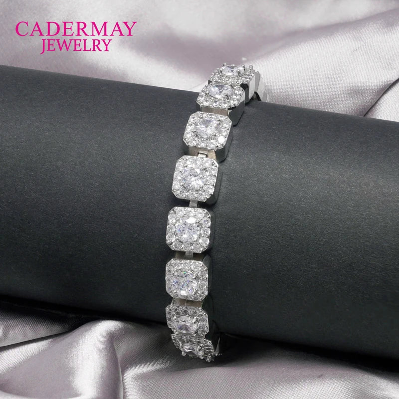 A silver-toned bracelet with six cushion-cut clear gemstones, displayed on a black cylindrical stand over a satin fabric background. The brand name 'CADERMAY JEWELRY' is visible in pink text in the top left corner.