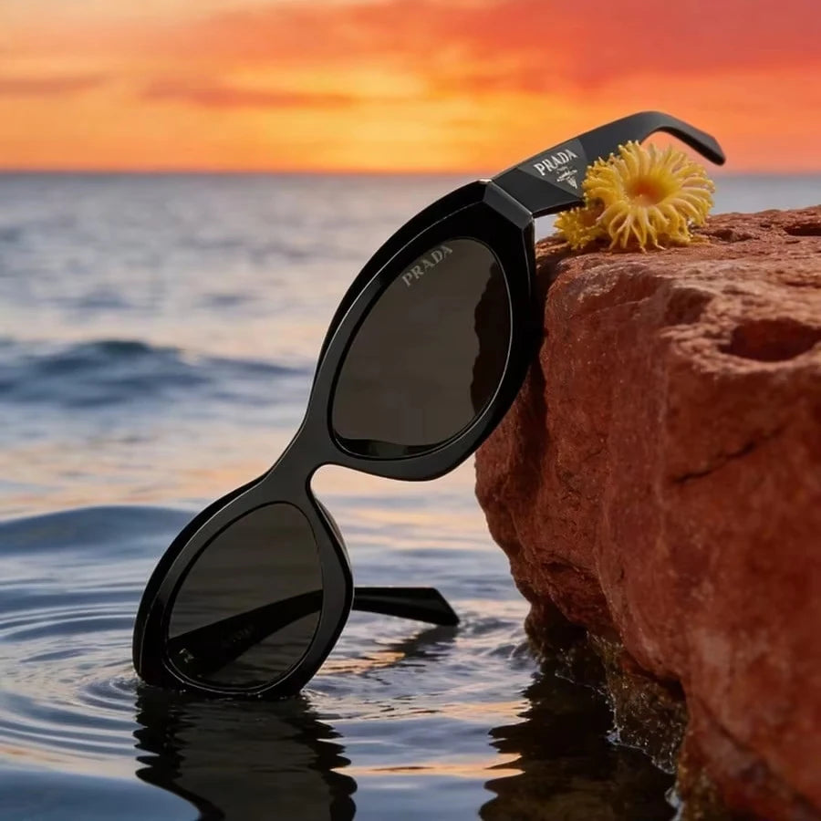 Black Prada sunglasses with dark lenses resting on the edge of a rocky shoreline at sunset, with ocean water in the foreground and a yellow flower on the rock. The Prada brand name is visible on the temple and lens.