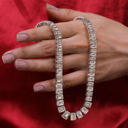 A silver tennis necklace with baguette-cut clear stones, draped over a hand with manicured nails, set against a rich red fabric background.