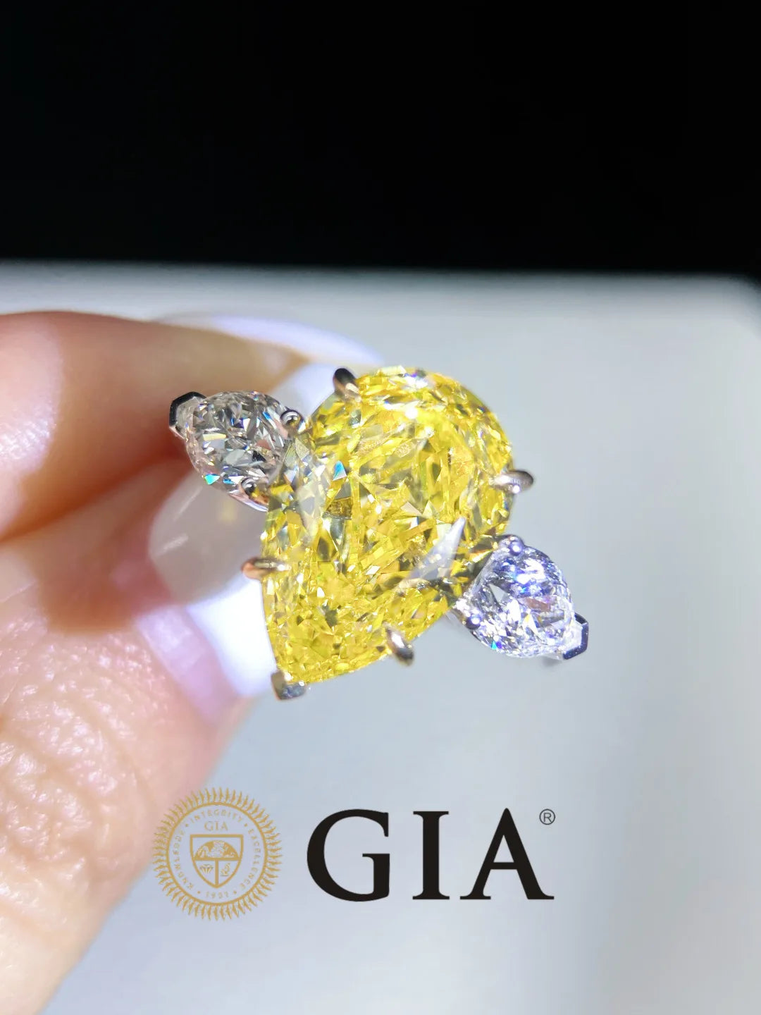 A close-up of a hand holding a GIA certified yellow pear-cut diamond ring with two small clear accent diamonds, set in a silver-colored metal, against a dark and light background with the GIA logo at the bottom.