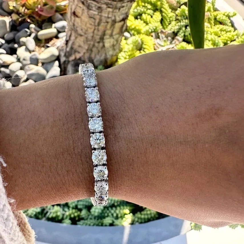 A close-up of a person's wrist wearing a silver tennis bracelet with a row of sparkling clear stones, set against a blurred outdoor background of green plants and rocks.