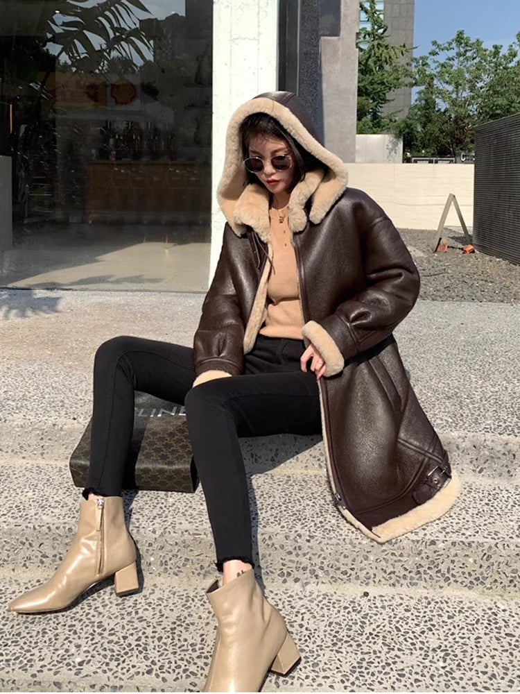 A woman sits on stone steps wearing a dark brown shearling-lined hooded jacket with beige fur trim, black pants, and beige ankle boots.