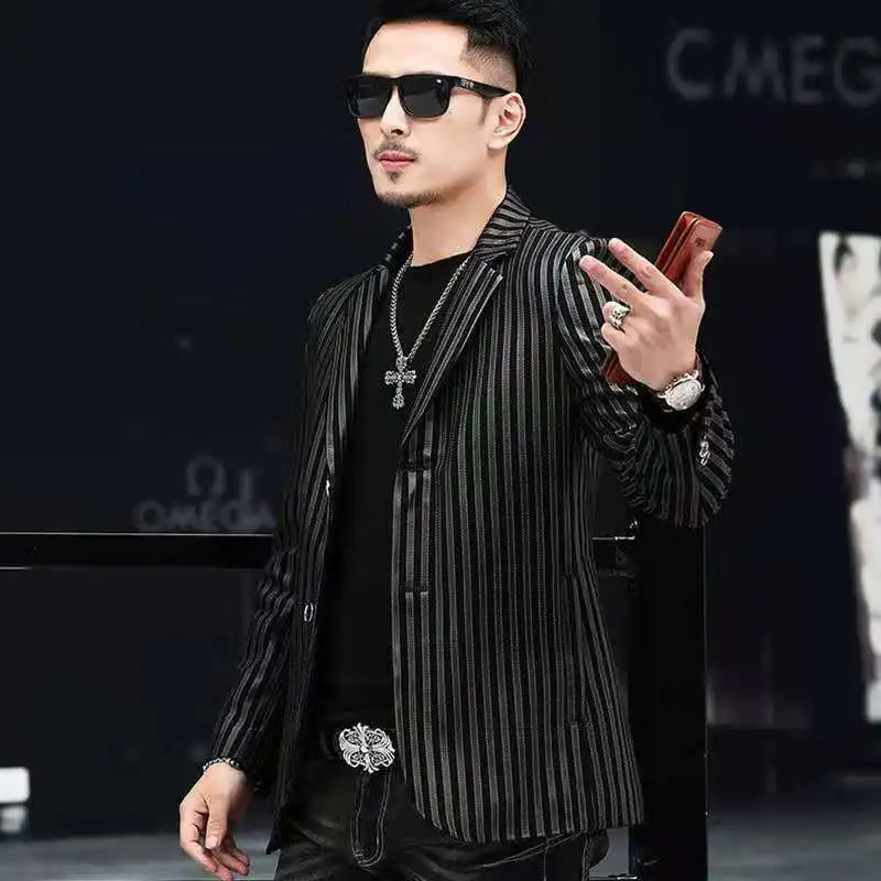 A man wearing a black and gray vertically striped blazer over a black shirt, accessorized with a silver cross necklace, sunglasses, and a watch, holding a brown phone case.