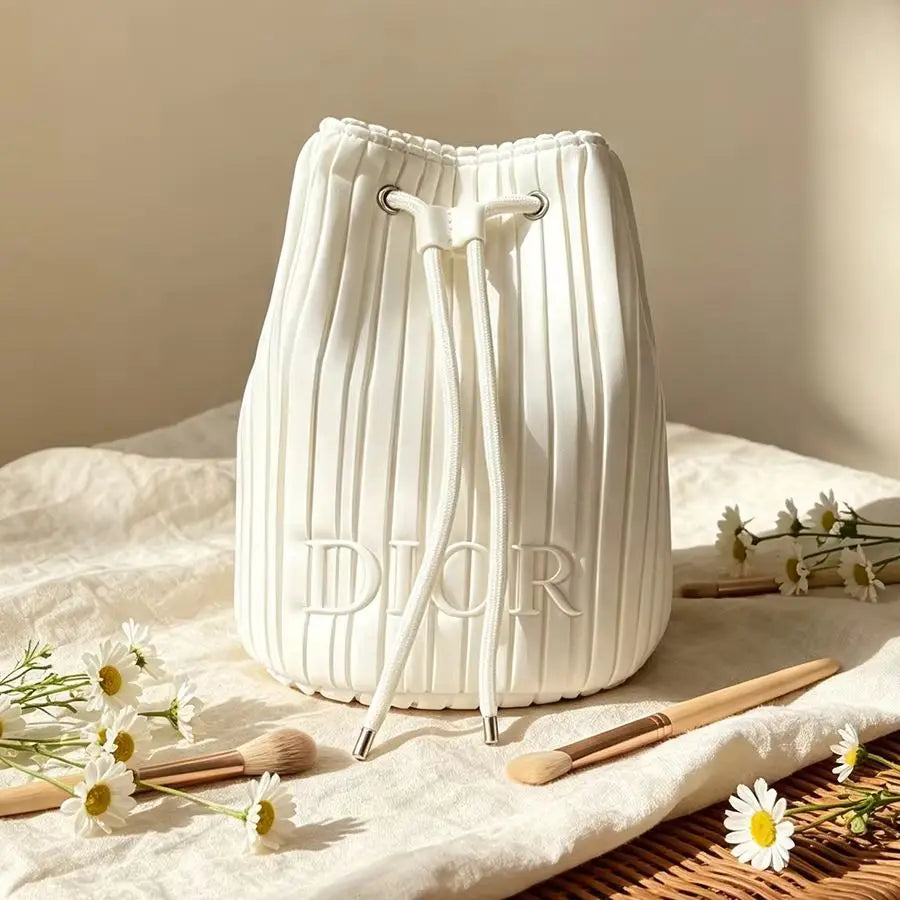 A white pleated bucket bag with a drawstring closure and embossed 'DIOR' lettering, styled on a cream fabric surface with small white flowers and wooden makeup brushes.