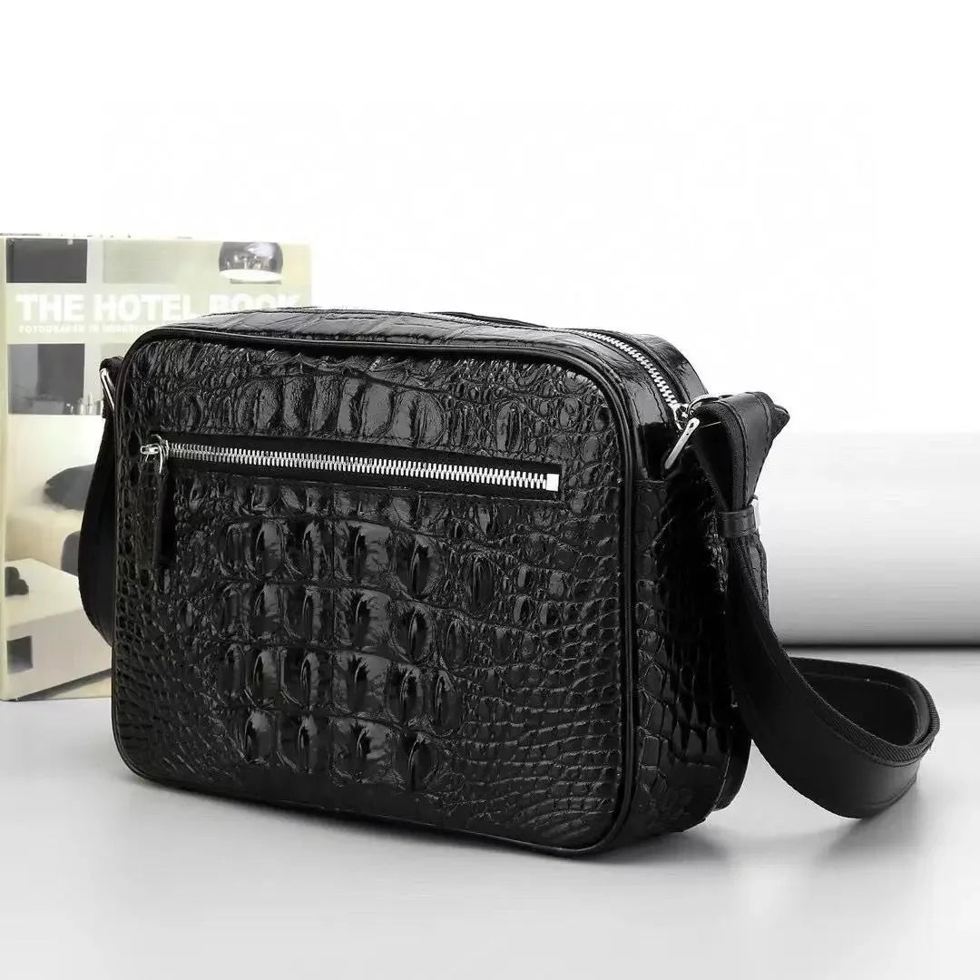 Black crocodile-embossed leather crossbody bag with a front zippered pocket and adjustable shoulder strap, shown on a light gray surface with a magazine in the background.