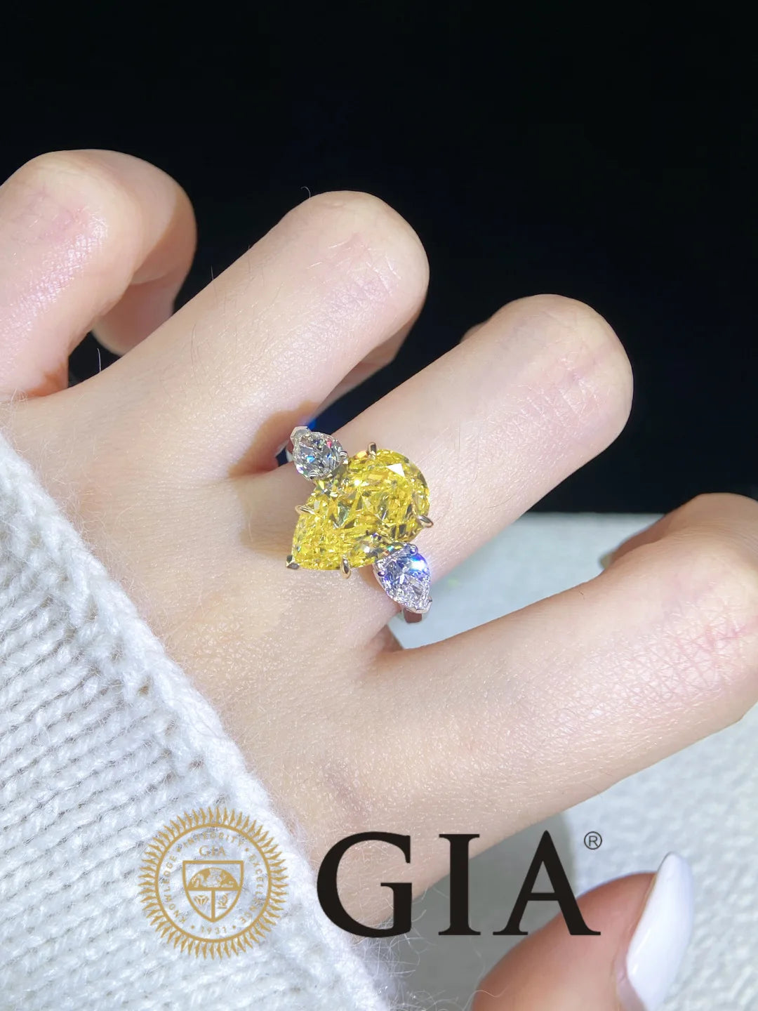 A close-up of a hand wearing a ring with a large yellow pear-cut center stone and smaller clear accent stones on a silver-toned band. The hand is dressed in a white knit sleeve, and a GIA logo watermark appears at the bottom.