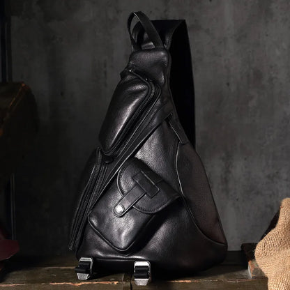 A black pebbled leather sling backpack with multiple zippered pockets and a single shoulder strap, displayed on a rustic wooden surface against a dark textured concrete wall.