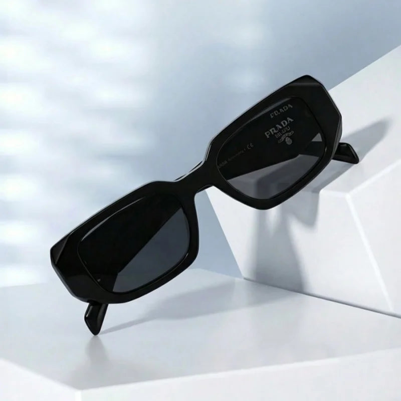 Prada black sunglasses with a geometric angular frame and dark lenses, displayed on a white geometric pedestal.