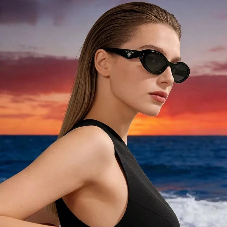 A woman wearing black Prada cat-eye sunglasses and a black swimsuit, posed against a dramatic sunset over the ocean.
