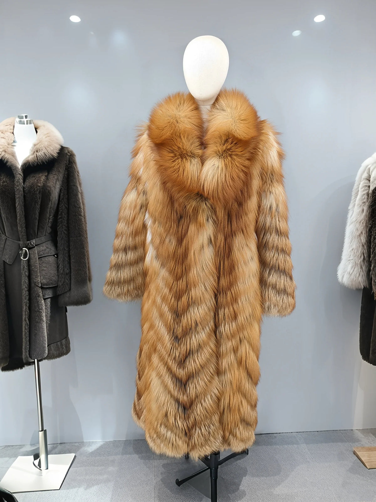 A long fur coat displayed on a mannequin in a showroom. The coat is tan and brown with a large shawl collar and striped fur panels.