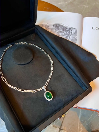 A silver-tone chain necklace with an oval green gemstone pendant, displayed inside an open black jewelry box on a black velvet cushion. The necklace is photographed from above, with an open book visible in the background.