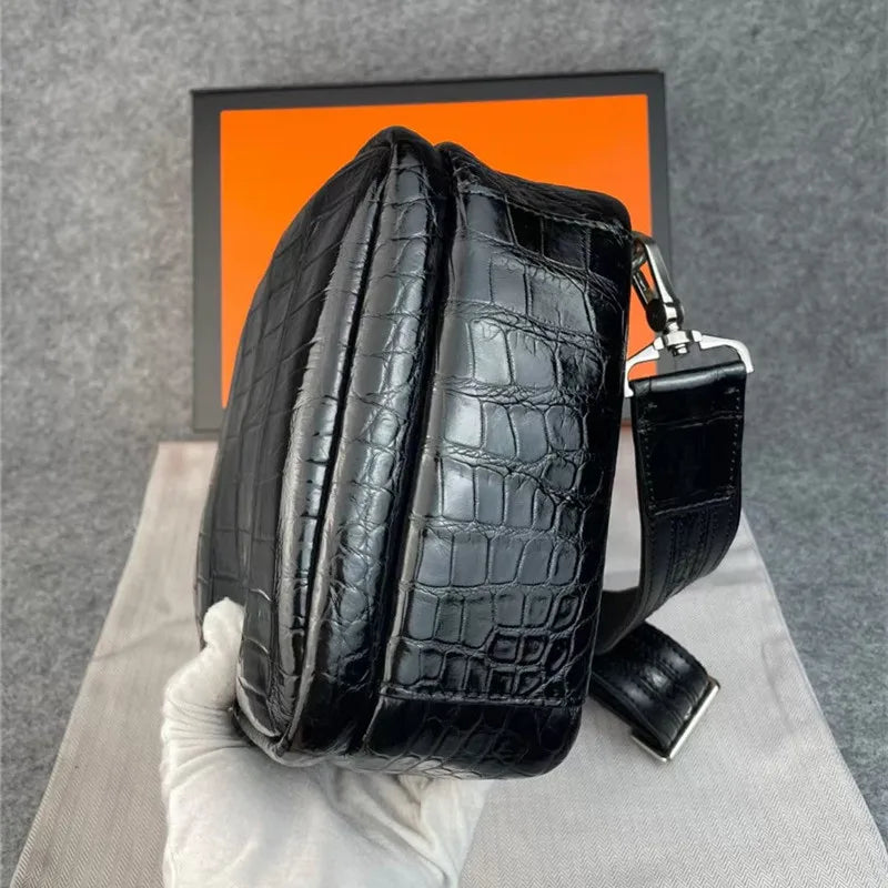 A glossy black crocodile-embossed leather shoulder bag held by a white gloved hand, with a bright orange box in the background.