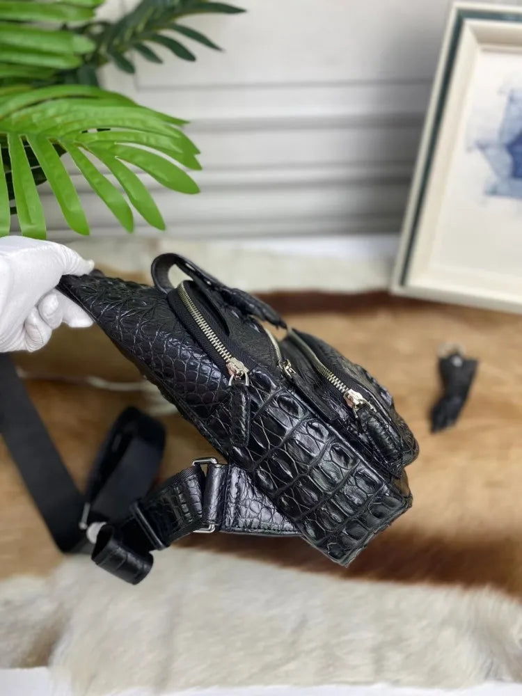 A black crocodile-embossed leather crossbody bag with gold-tone hardware, held by a white-gloved hand. The bag features multiple zippered compartments and a matching black shoulder strap.
