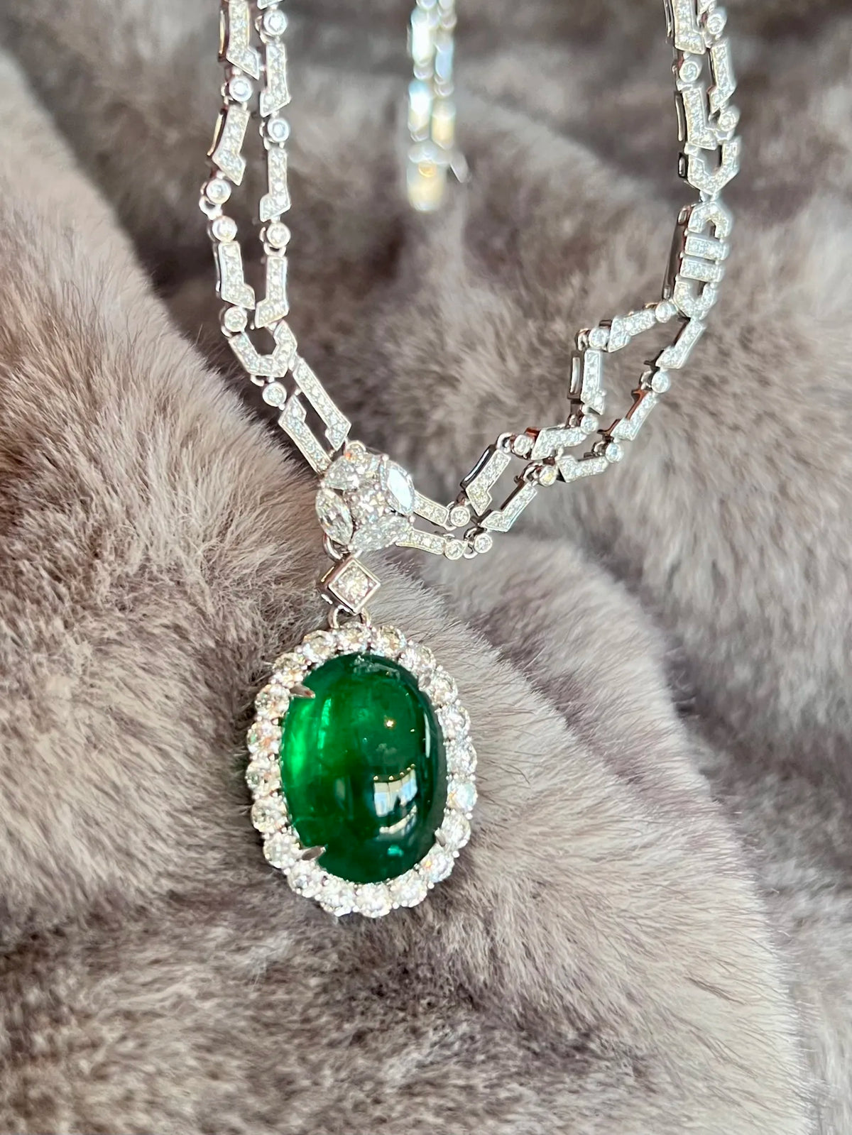 An oval emerald-cut green gemstone pendant necklace with a silver-toned halo and chain, displayed on a textured gray faux fur surface.
