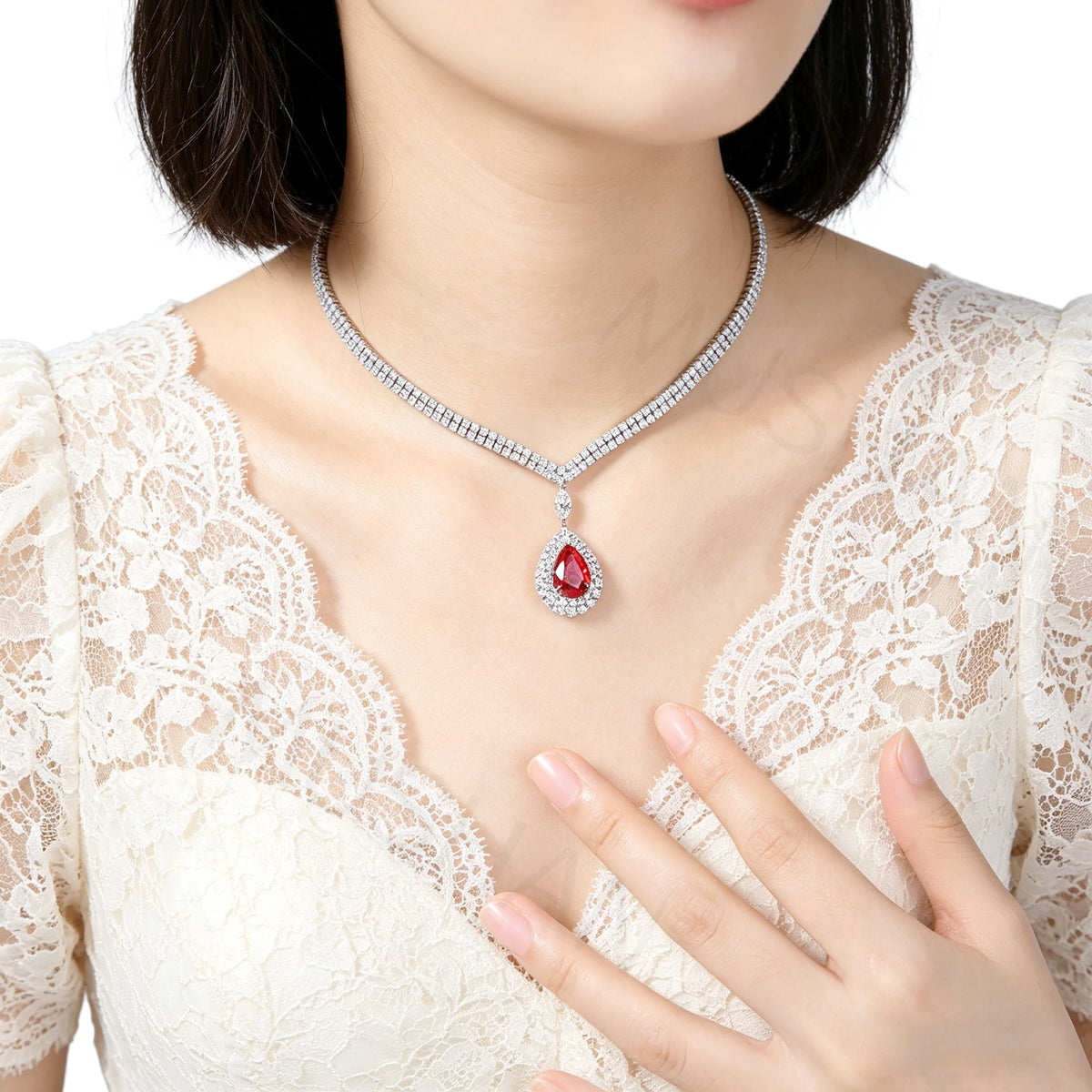 A close-up of a model wearing a silver-toned diamond-studded necklace with a teardrop-shaped red gemstone pendant, paired with a cream lace top.