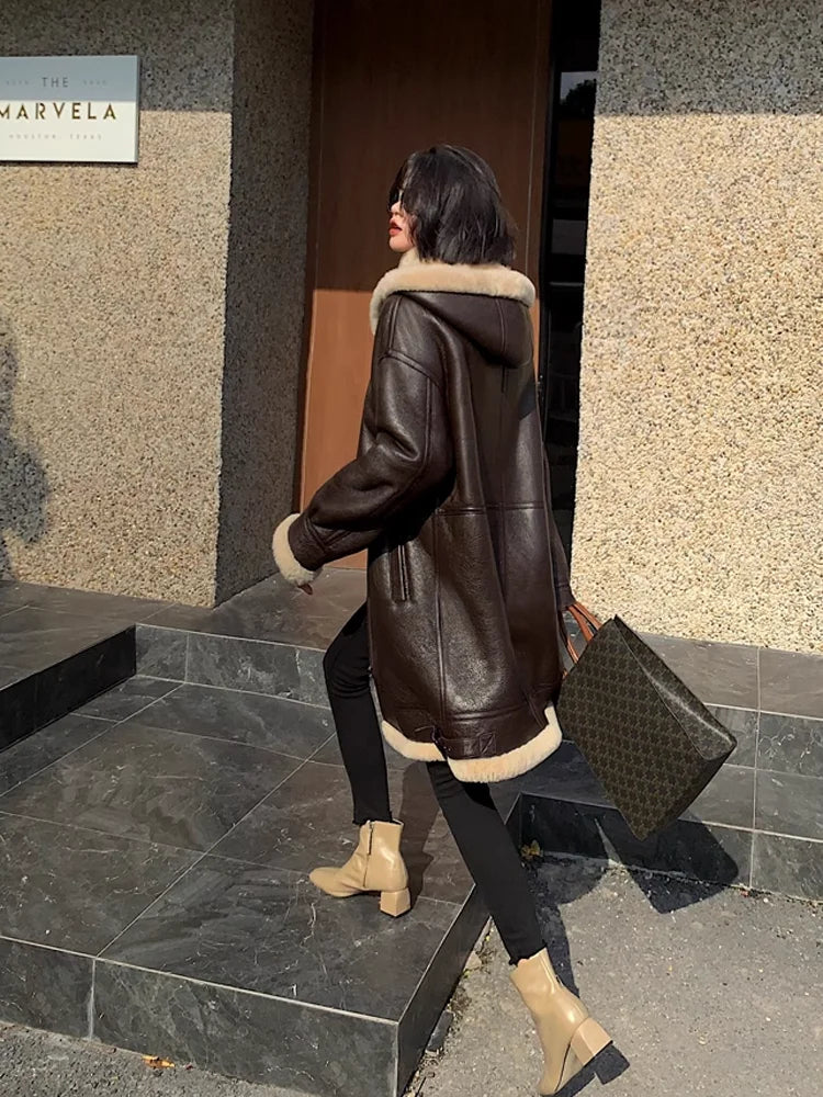 A woman wearing a dark brown faux-leather hooded coat with beige shearling trim walks down stone steps, carrying a dark brown patterned tote bag.