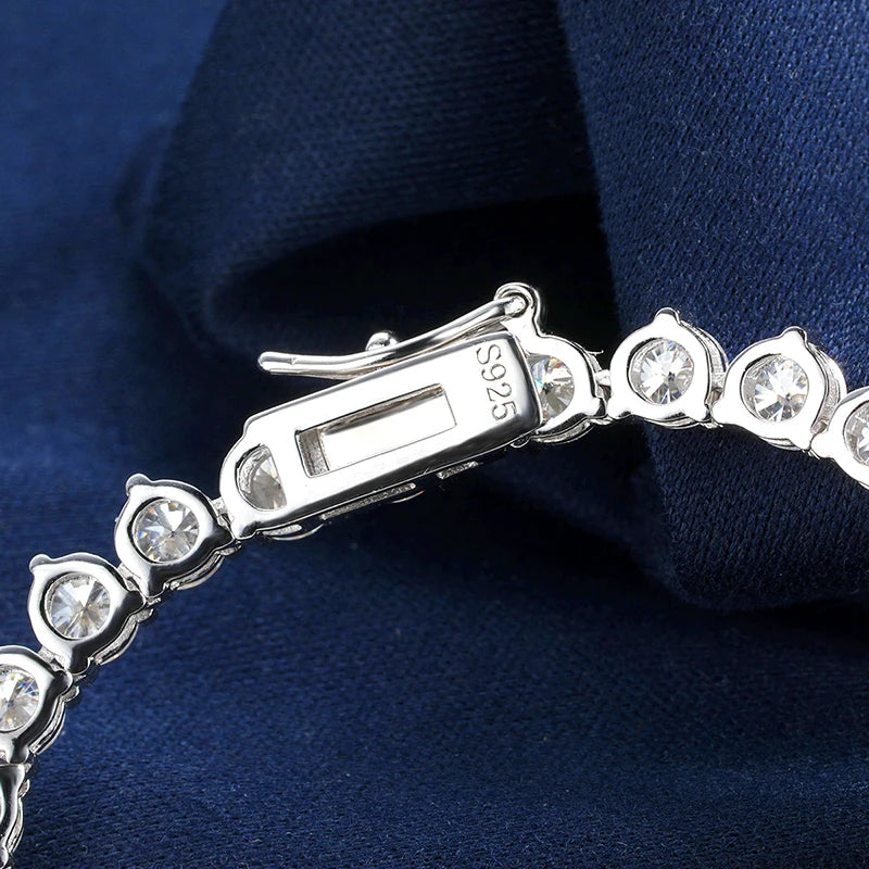Close-up of a silver tennis bracelet with clear round-cut stones and a clasp stamped S925, resting on dark navy blue fabric.