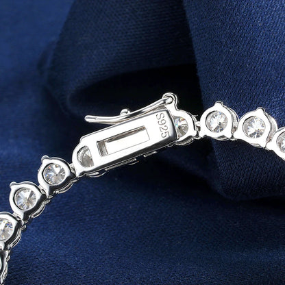 Close-up of a silver tennis bracelet with clear round-cut stones and a clasp stamped S925, resting on dark navy blue fabric.